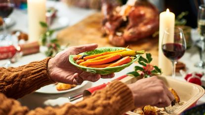 table with vegetables and serving dish of roasted carrots, roast turkey resting in background, lit candles and red wine