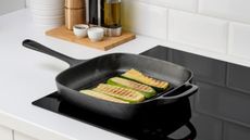 a square cast iron griddle pan on a black electric stove top, frying slices of courgette