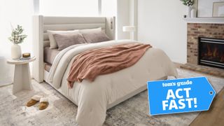 Sleep Number bed dressed with beige bedding with beige headboard a bedside table with plant to the left, fireplace to the right and brown slippers next to bed with Tom's Guide 'Act Fast' logo