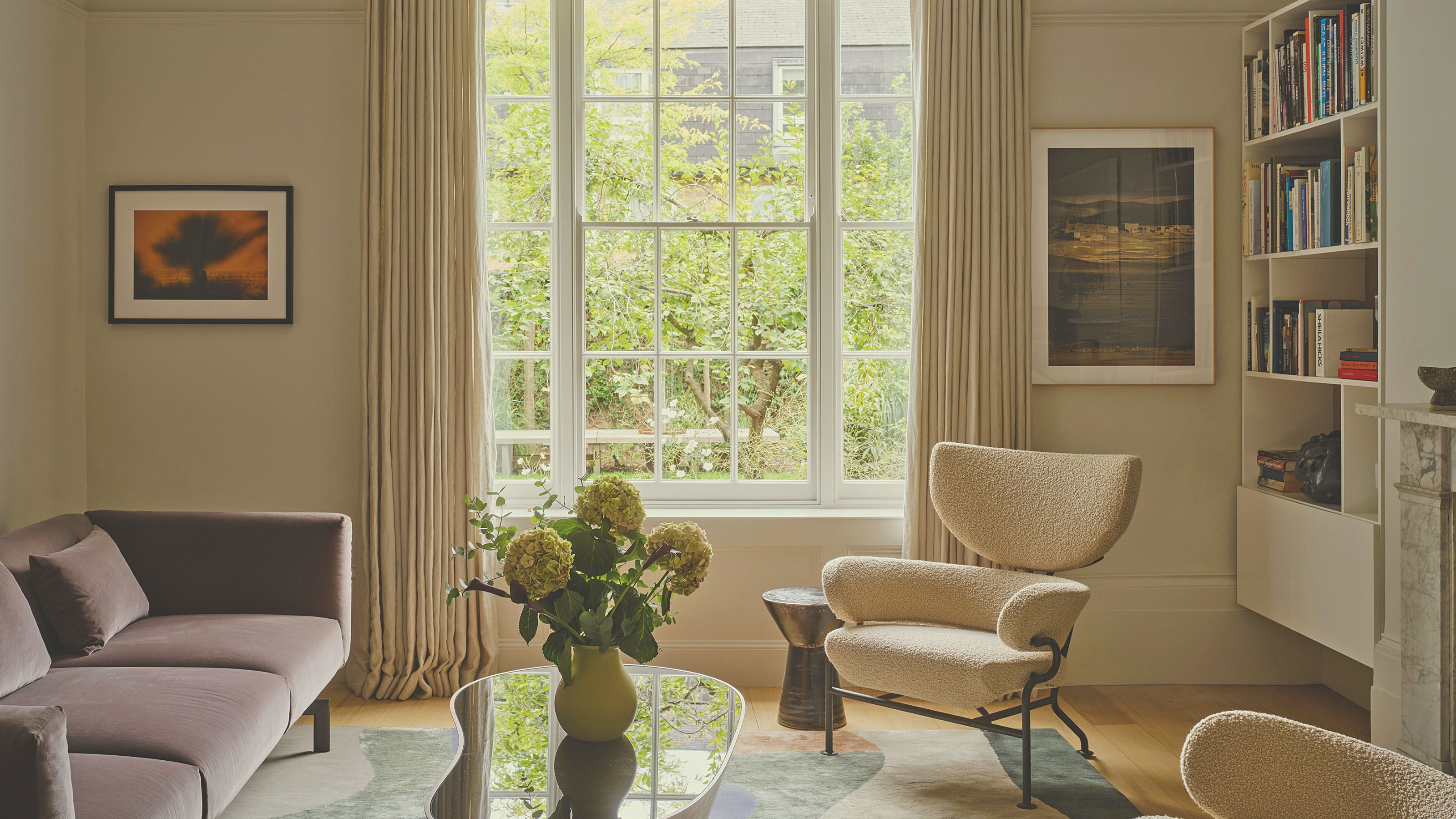 A sunlit contemporary living room by Atelier Como featuring a mauve velvet sofa, a cream boucl&amp;eacute; lounge chair, and a large window overlooking a green garden.
