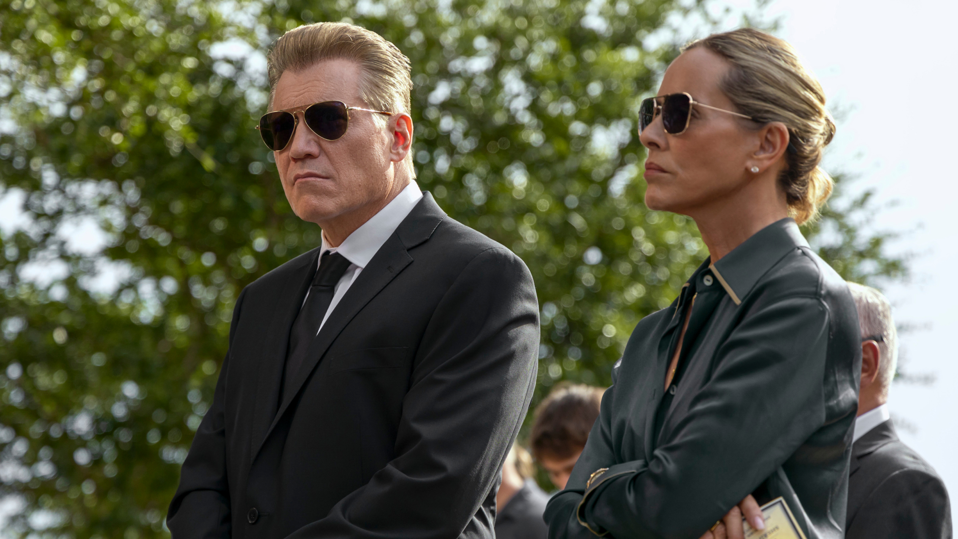 Holt McCallany and Maria Bello as the buckleys standing at a funeral with sunglasses on in the waterfront
