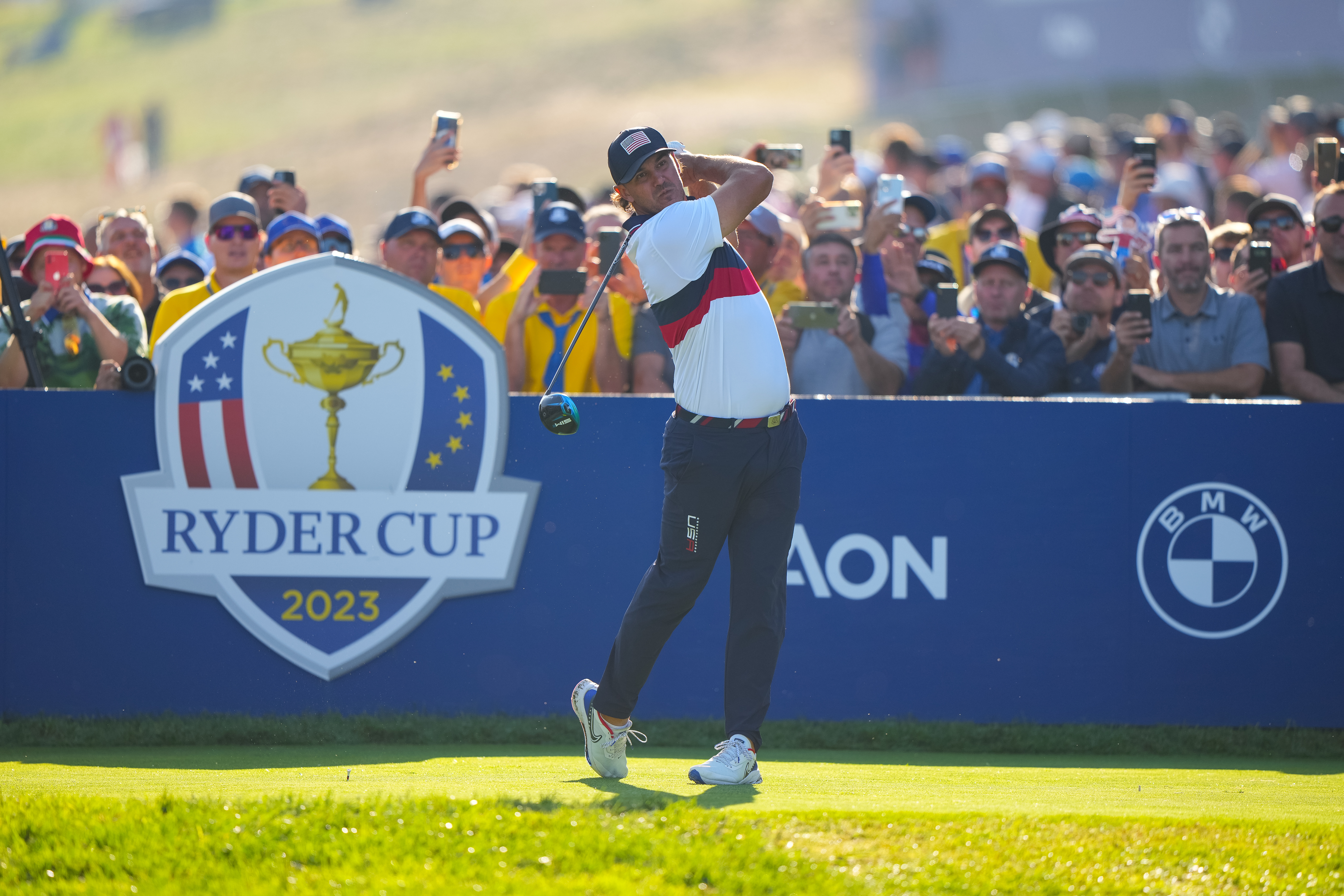 Brooks Koepka hits a tee shot in front of a Ryder Cup board