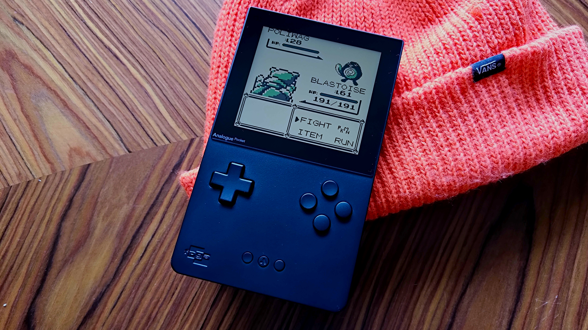 Analogue Pocket lying on orange Vans beanie with Pokemon Blue battle on screen featuring Blastoise vs Poliwag.