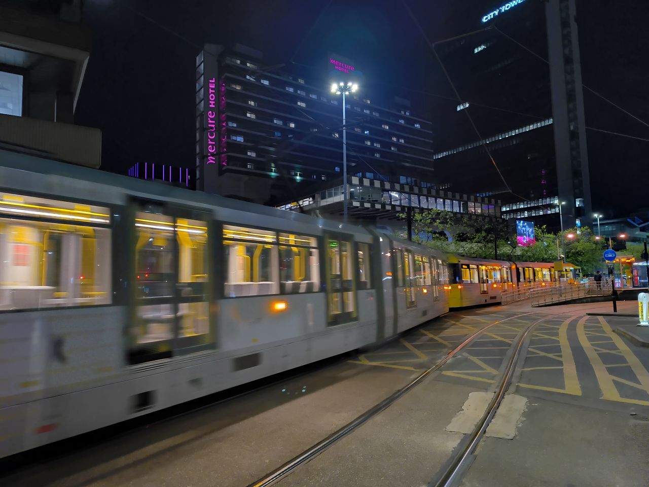 A light rail tram passing through a city center at night with illuminated carriages.