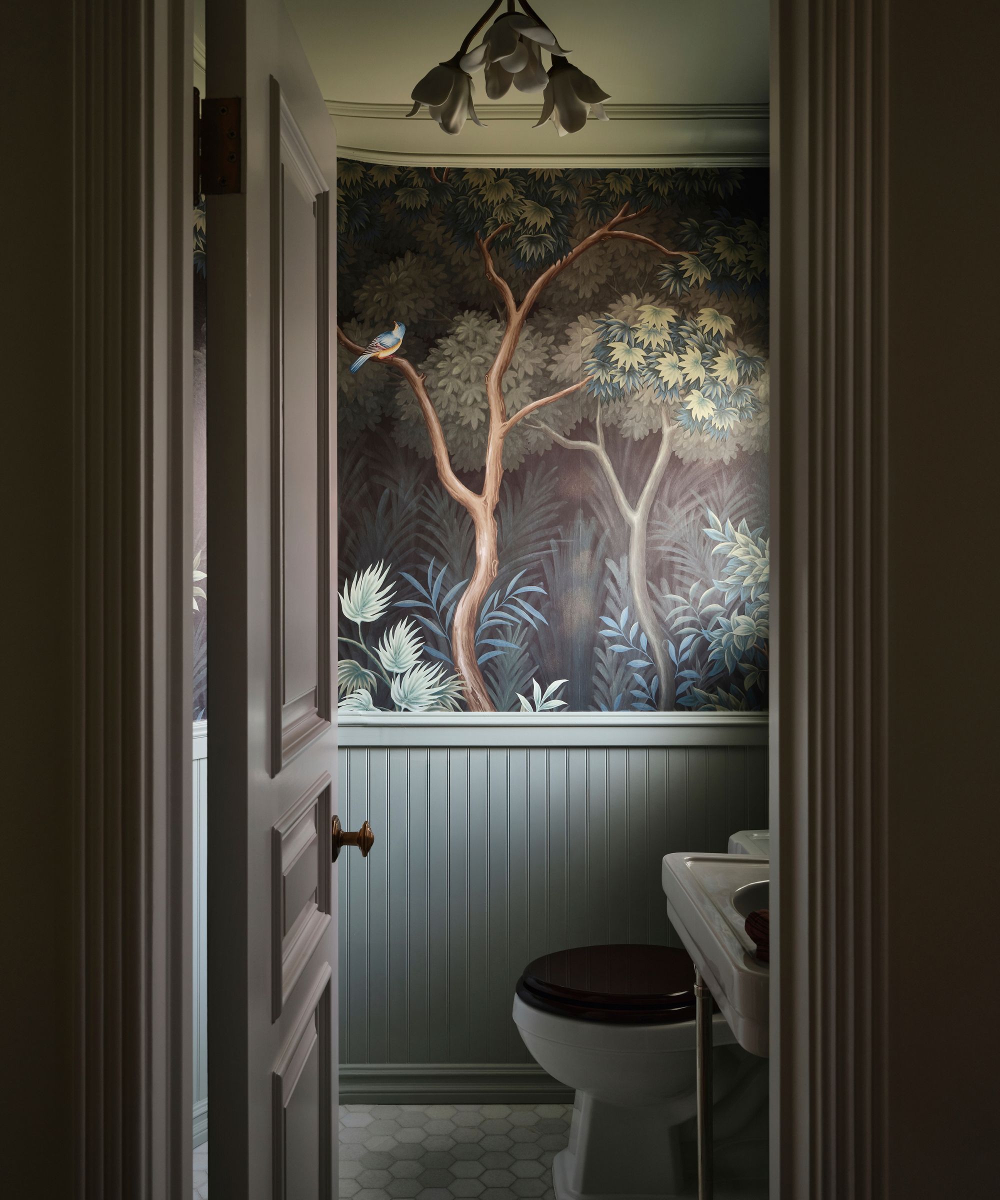 View through a doorway of a powder room with dark, elaborate forest-mural wallpaper, light green wainscoting, a pedestal sink, and a toilet