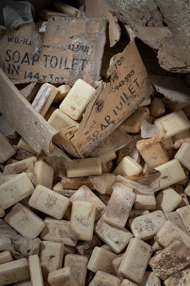 A heap of unused bars of soap stamped with a crown, spilling out of torn cardboard boxes printed with "SOAP TOILET" and stock numbers, covered in dust