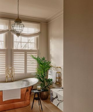 neutral quiet luxury bathroom with a freestanding copper bath and window shutters with a roman blind