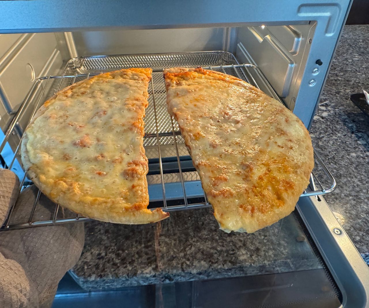 The Typhur Sync Air Fryer Oven on a granite work top, open, pulling a cheesy flatbread out on a wire tray.