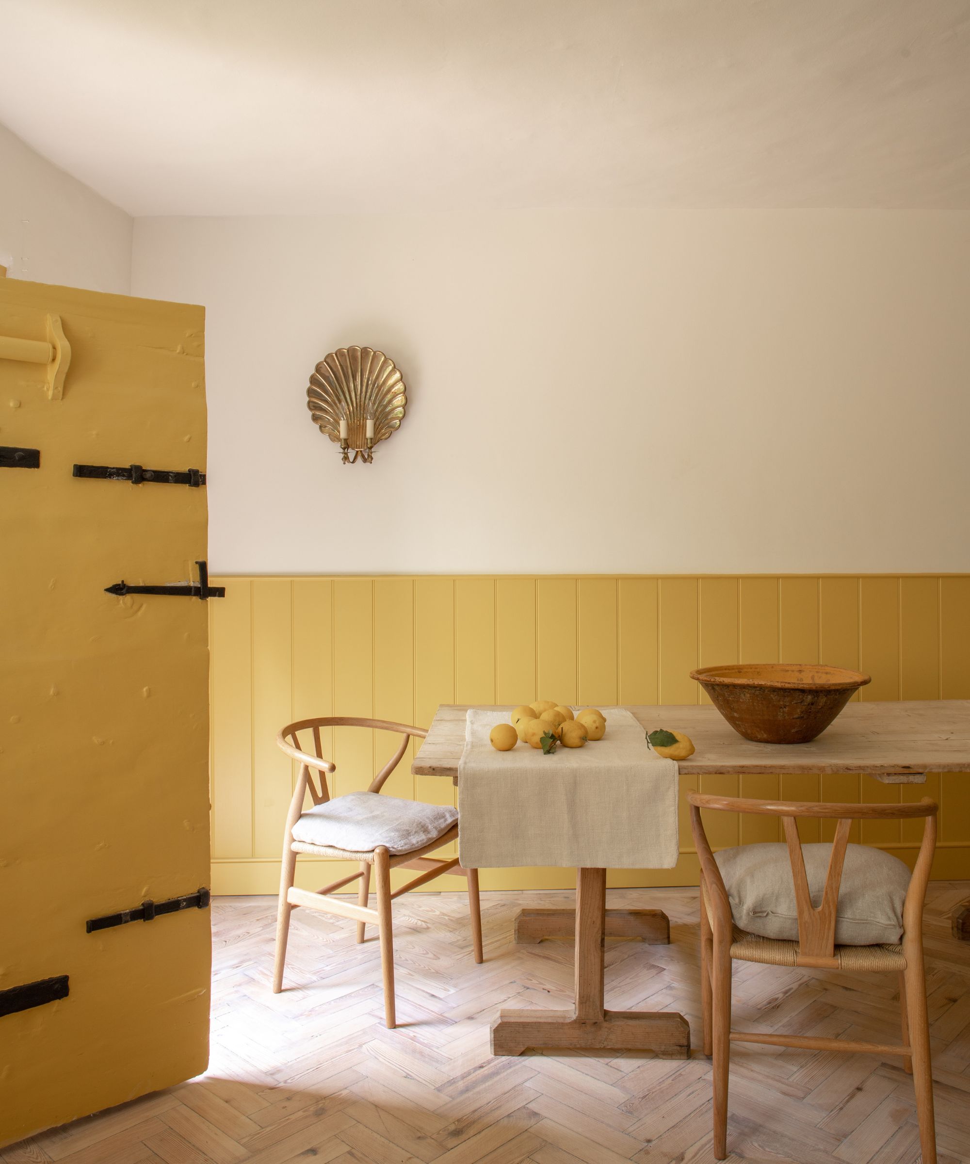 Farmhouse dining room with yellow painted wall panelling, a pale oak dining table, Scandinavian-style chairs, a yellow painted front door, and wooden parquet floors, with a gold shell sconce above.