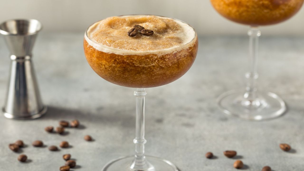 A glass of Frozen Espresso Martini, topped with coffee beans next to a stainless steel jigger
