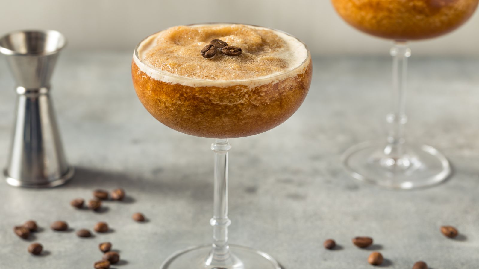 A glass of Frozen Espresso Martini, topped with coffee beans next to a stainless steel jigger