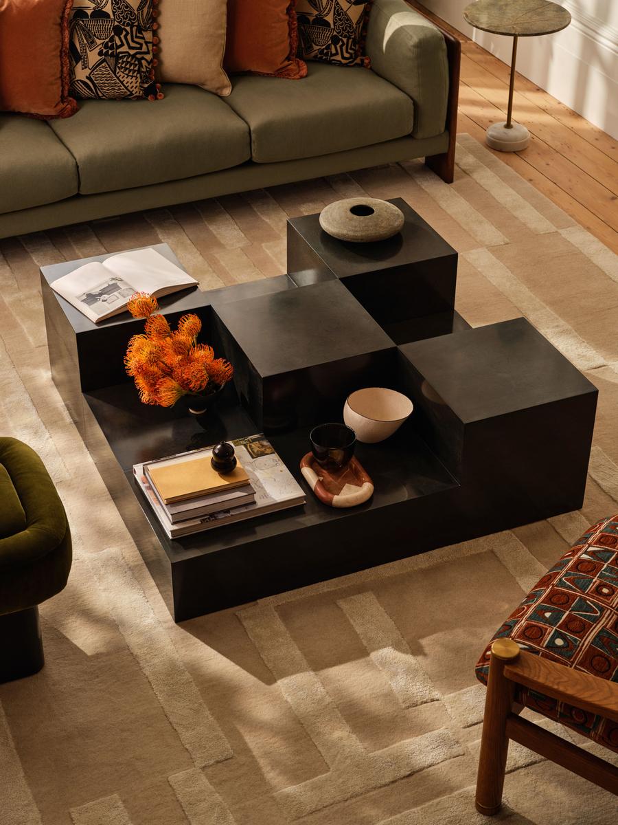Tomasso Coffee Table in black arranged in the center of a beige, low-pile rug. There is part of a sofa and two chairs in view.