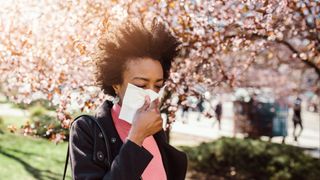 black woman walking through a city park with blooming cherry trees in the background as she blows her nose into a tissue