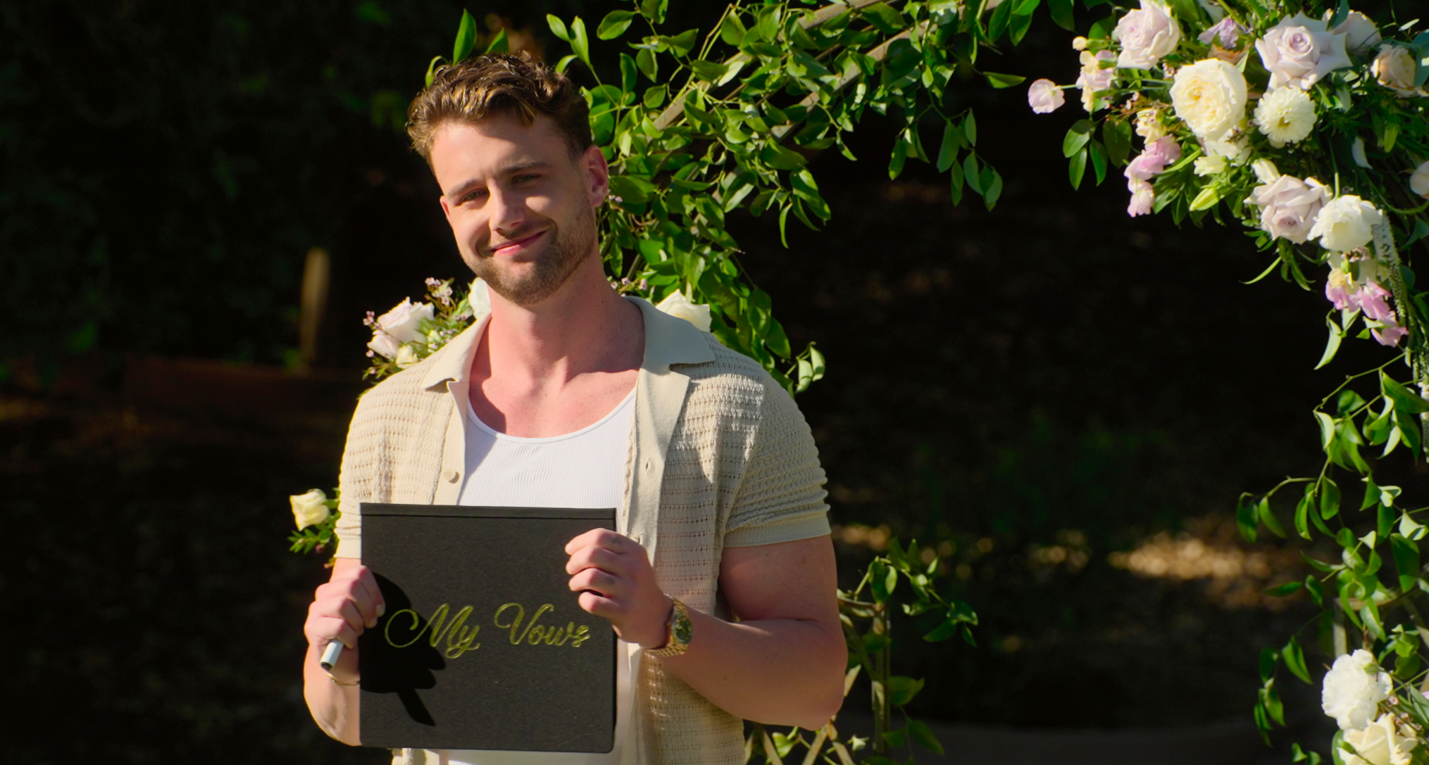 reality star harry jowsey stands in front of a floral display holding a sign that says my vows in a still from the show let's marry harry