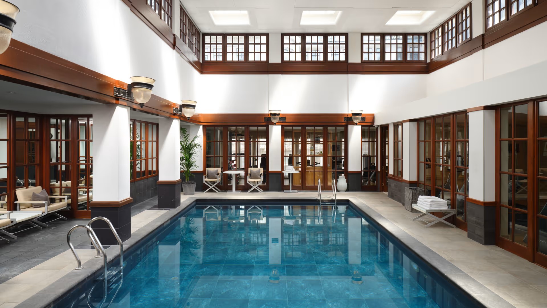 An indoor pool with white-washed walls, wooden detailing, stone floors, beige beach chairs in leather, and a beautiful, rock-like pool floor.