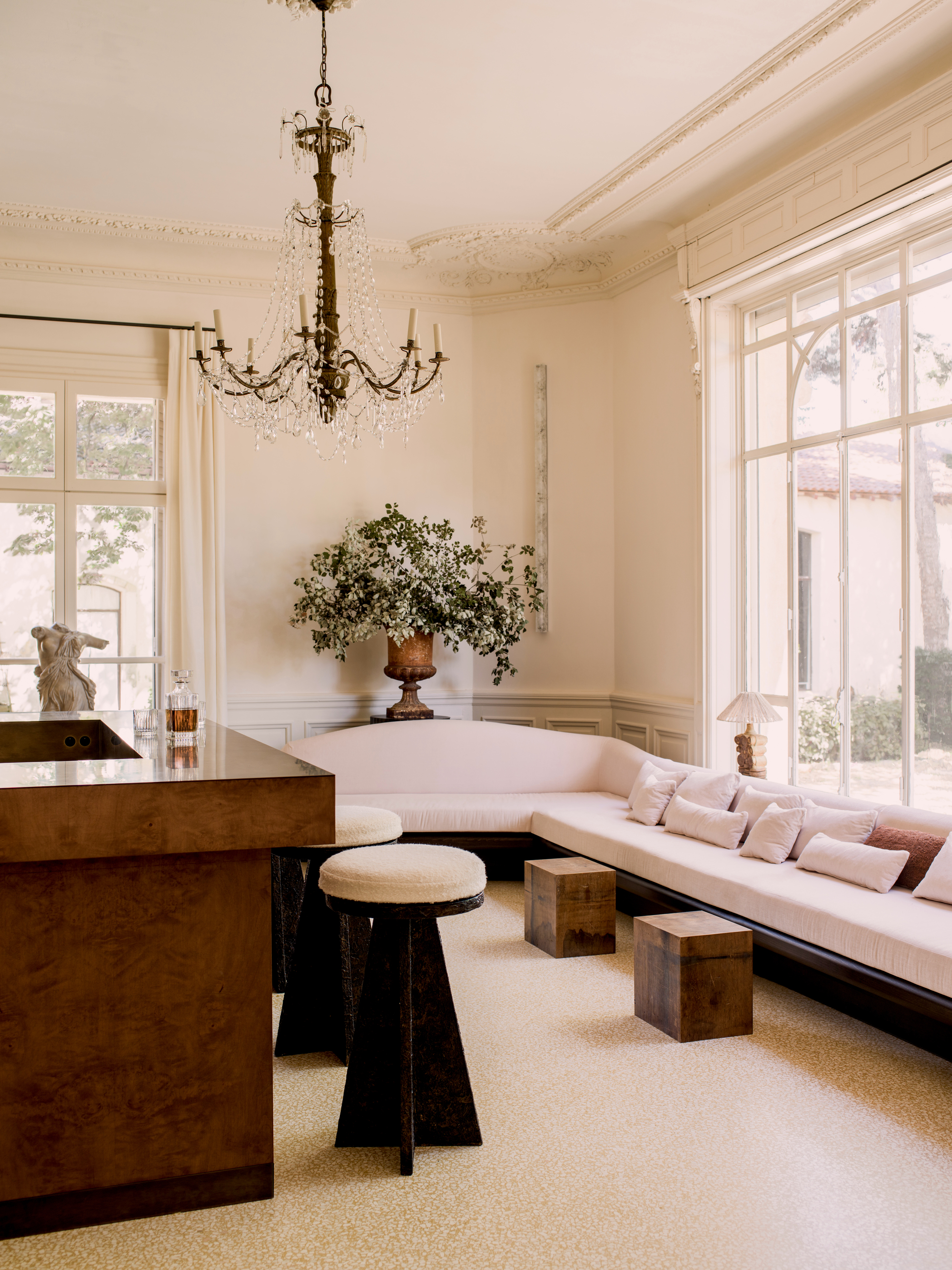 Image of a large room with crown molding and large windows. There is a large sectional sofa that is blush pink with a curved back. There is a burl wood bar and modern stools.