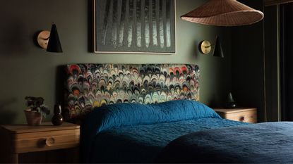 green bedroom with marbled print fabric headboard, navy blue bedding, timber side table with pot plant, wall sconces, rattan pendant light, and artwork over bed