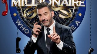 Jimmy Kimmel pretending to talk on the phone while wearing a suit and rocking a side part for Who Wants To Be A Millionaire.