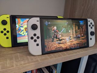 Två stycken Nintendo Switch OLED står uppställda och kör ett varsitt spel, där den i bakgrunden har gula JoyCons och den längst fram har vita.