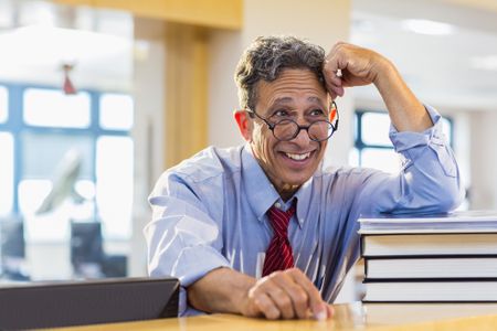 A man with greying hair wearing a button-up shirt with a tie and glasses laughs standing at a library circulation desk.