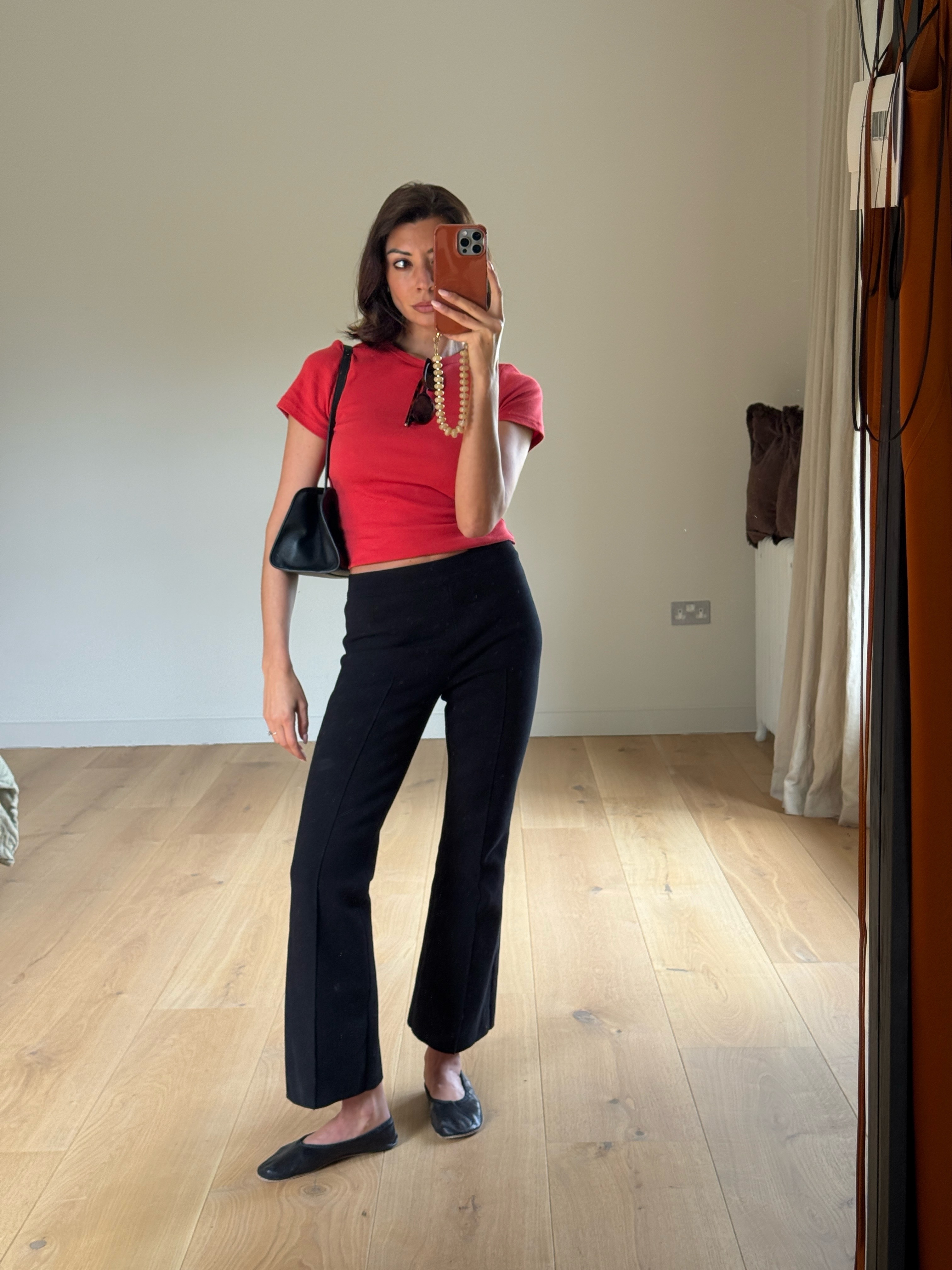 Fashion person Marianne Smith wears June Outfit Ideas 2026. She wears a red crop top, black kick flare trousers and minimal black ballet flats. She has The Ro René bag under her arm.
