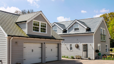 A home with a double garage and shingle roof. The exterior is clad in dove-gray wood and the windows and doors are white with black hardware