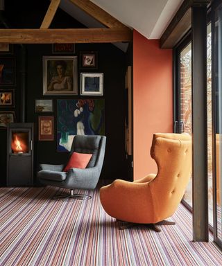 Living room with vaulted ceiling, wooden beams, orange painted feature wall, orange lounge chair, striped colorful carpet, large floor to ceiling windows, gray armchair, wood burning stove, gallery wall