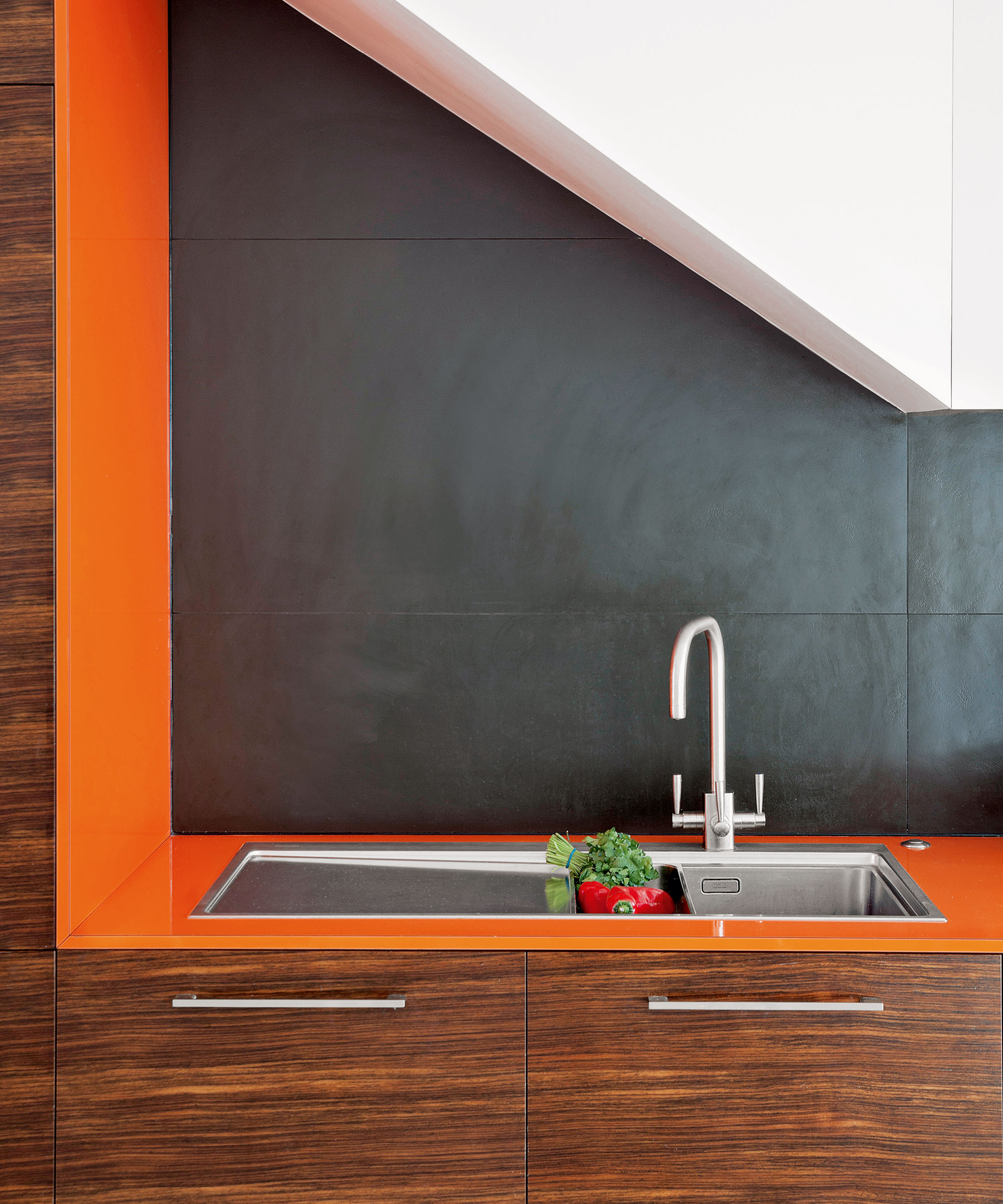 A close-up shot of a modern kitchen with a 1970s retro aesthetic, featuring a vibrant orange countertop and matching vertical trim. A stainless steel undermount sink with a gooseneck faucet is set into the counter, holding fresh red peppers and herbs. The cabinetry below is made of a dark, horizontally grained wood, while the backsplash consists of large, matte black tiles. An angled white architectural element is visible at the top of the frame.