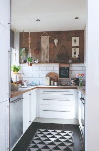 small u-shaped kitchen with white units, oak worktops, and wood panelling on upper third of wall with macrame wall hangings