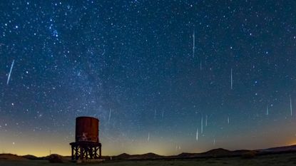 A dark blue night sky is streaked with white meteors moving through space with a rusty cylindrical water tower below.