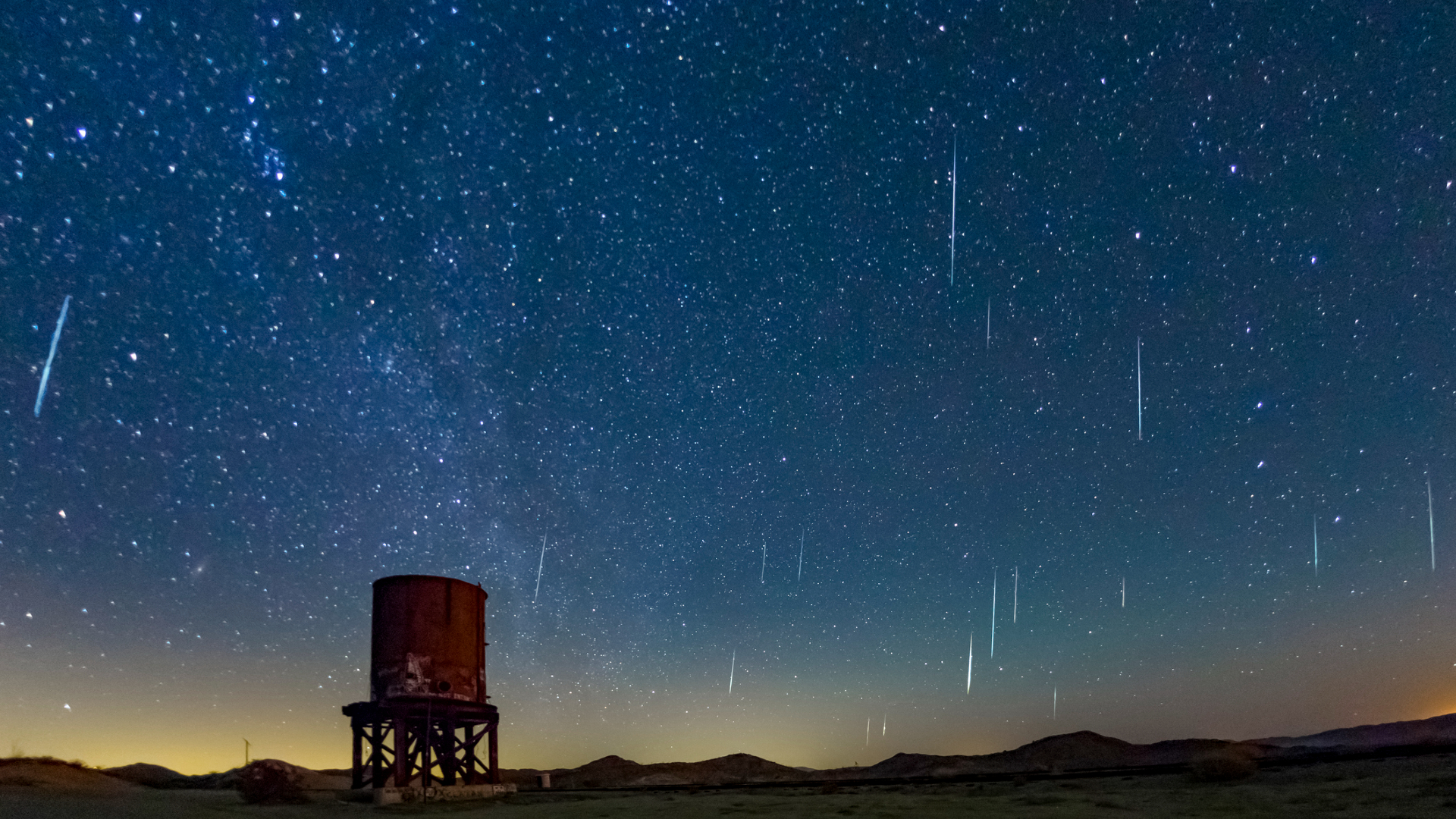 A dark blue night sky is streaked with white meteors moving through space with a rusty cylindrical water tower below.