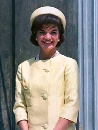 Jackie Kennedy smiling in the sunshine on a visit to Paris wearing a yellow suit, pearl necklace, earrings and a yellow pillbox hat