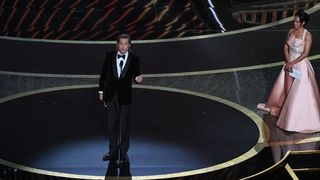 Brad Pitt delivers his acceptance speech at the Oscars as Regina King looks on from the side of the stage.