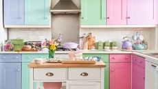 Pastel kitchen ideas are so cute. Here is one of these - a kitchen with lilac, mint green, pink and purple cabinets, a white counter with colorful decor on, and a white kitchen island with flowers on top