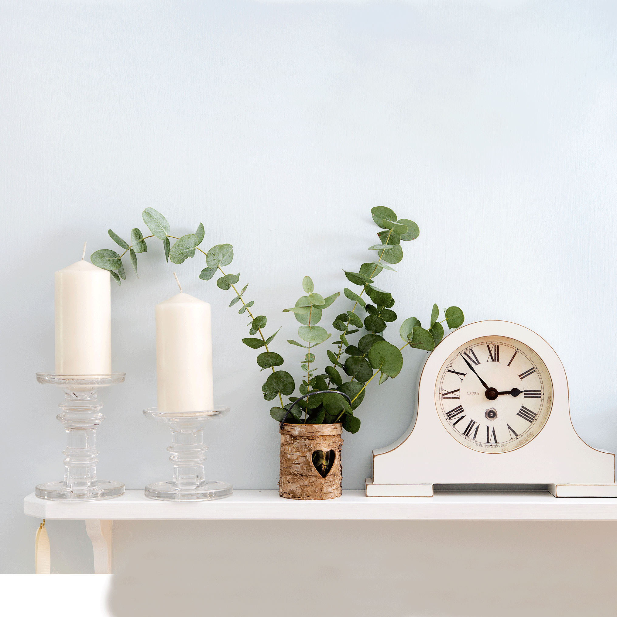 A white shelf supporting a pair of candles, a green leafy plant in a wooden pot, and a white analogue clock.