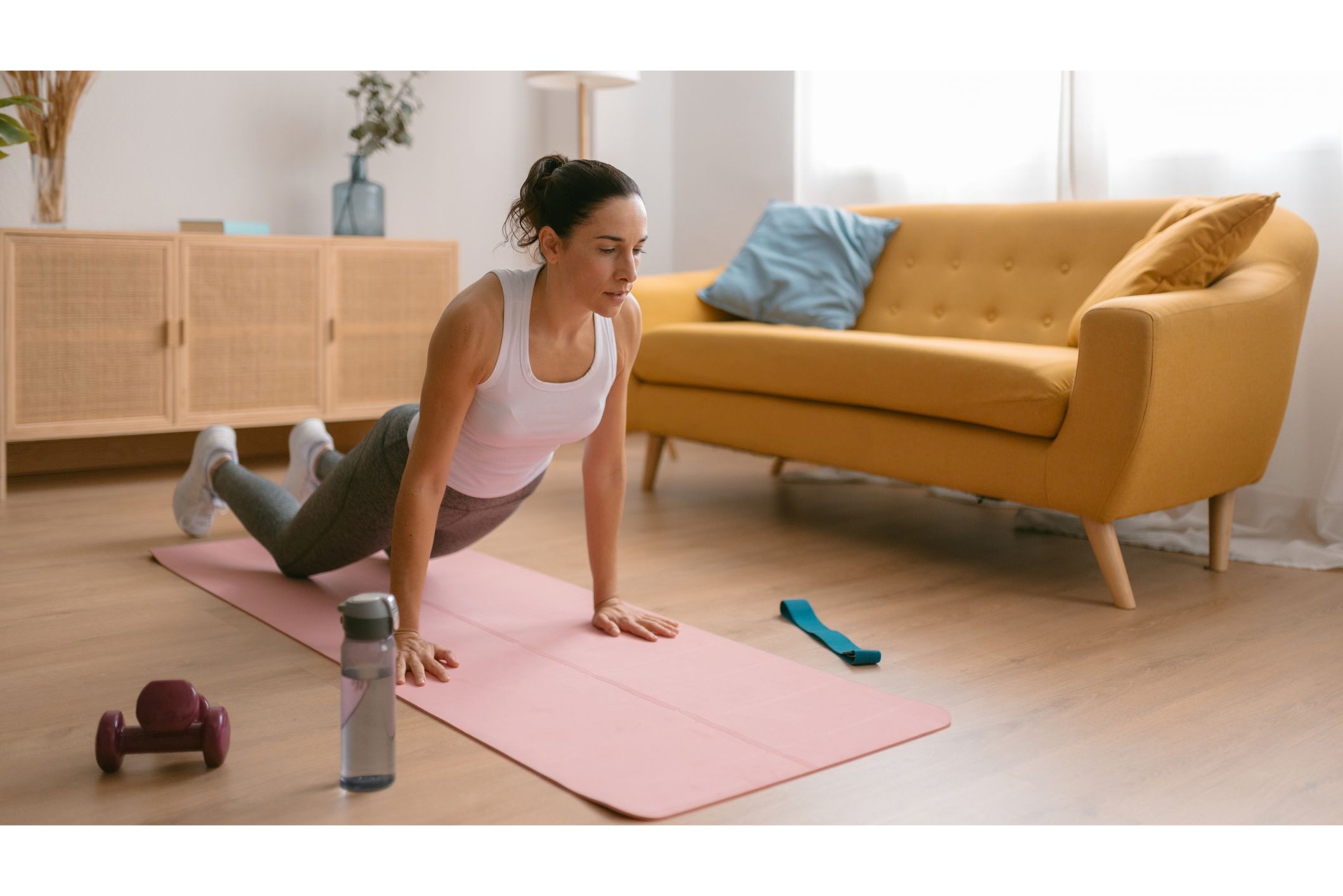 Woman exercising on a mat next to a couch with a resistance band, light dumbbells and a water bottle surrounding her