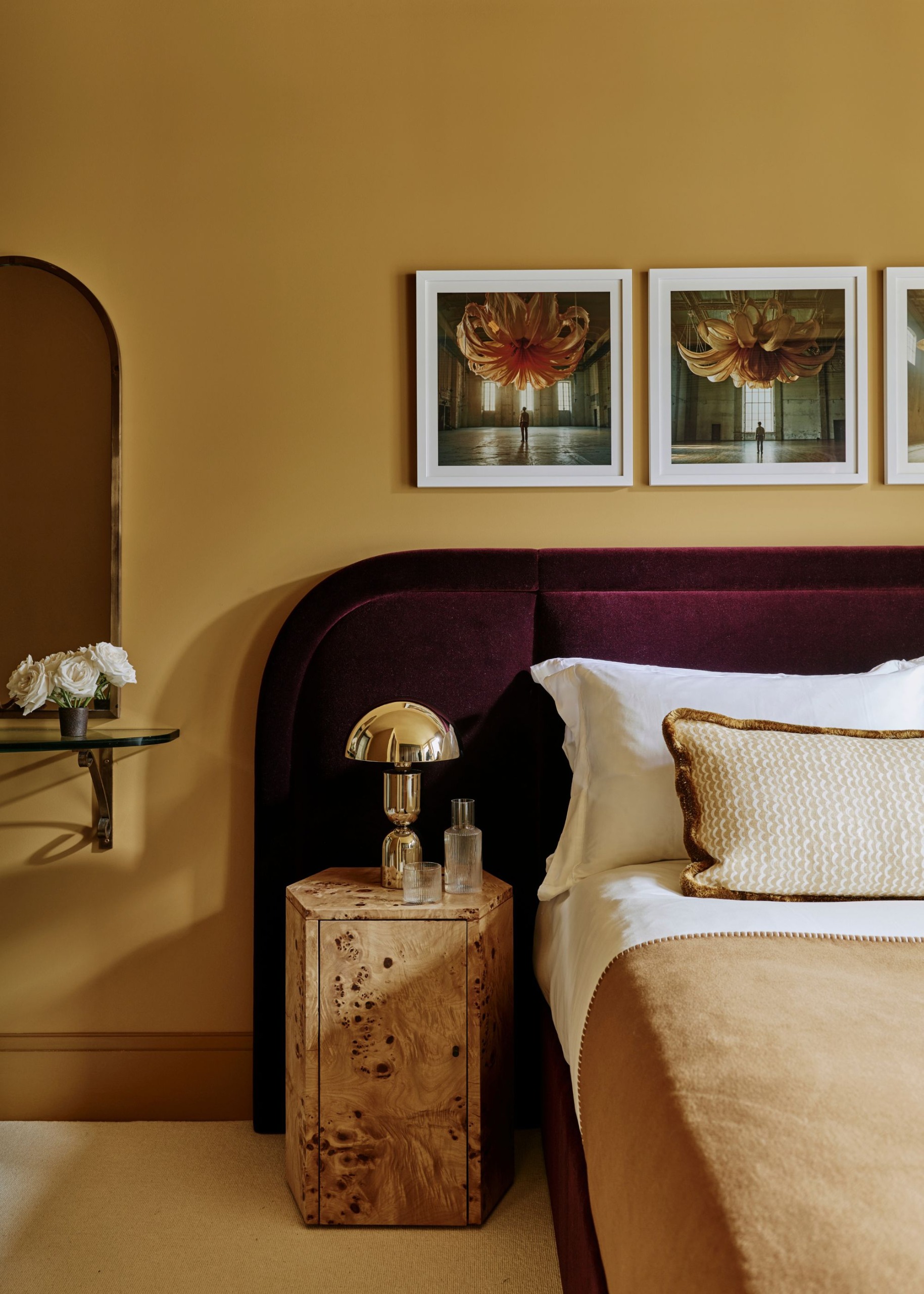 A cozy, modern bedroom with ochre walls, an elongated dark plum upholstered headboard, a burl wood nightstand, crisp white and caramel bedding, and three artworks on the wall.