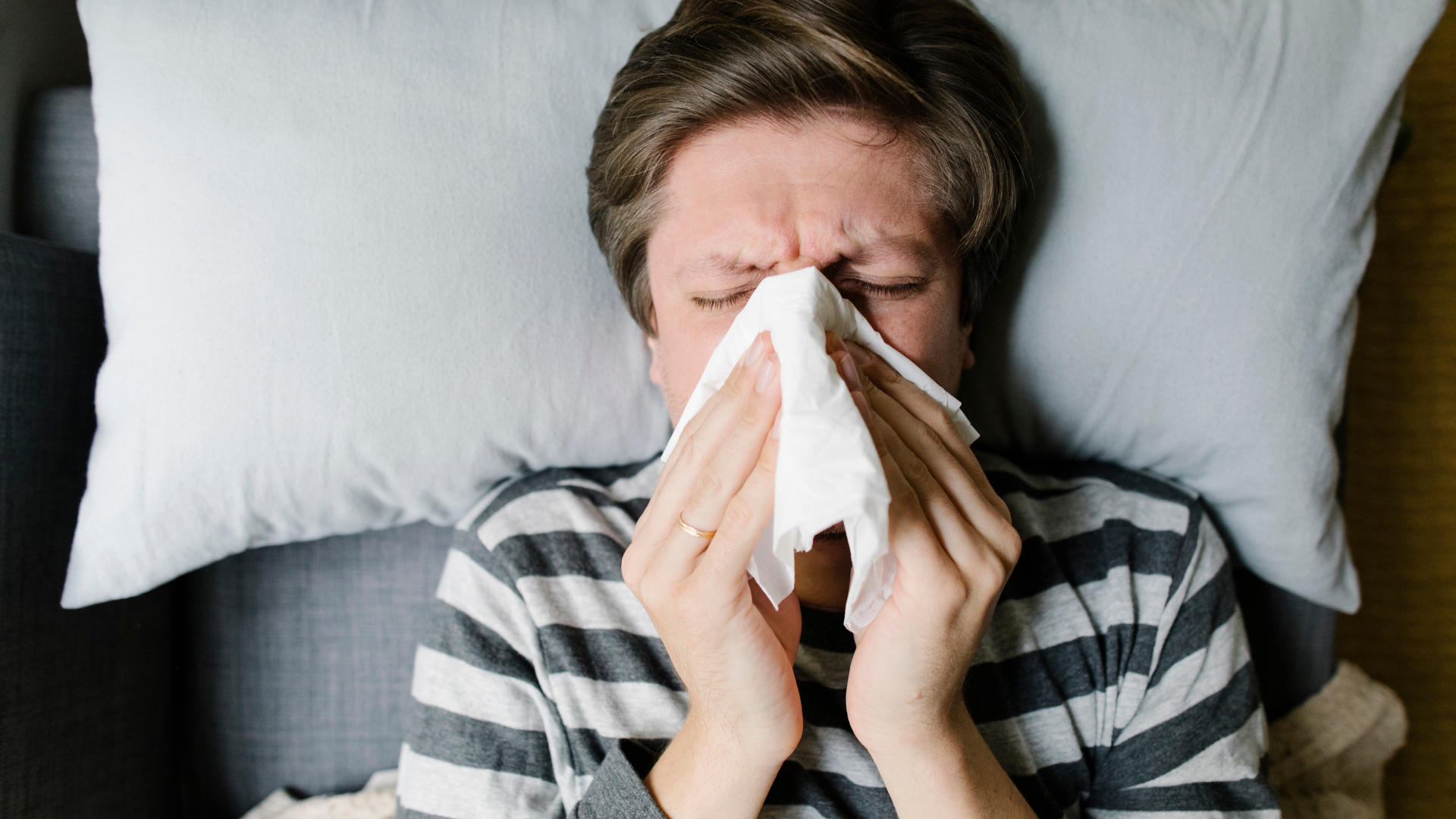 A man lies on his back in bed blowing his nose with a white tissue.