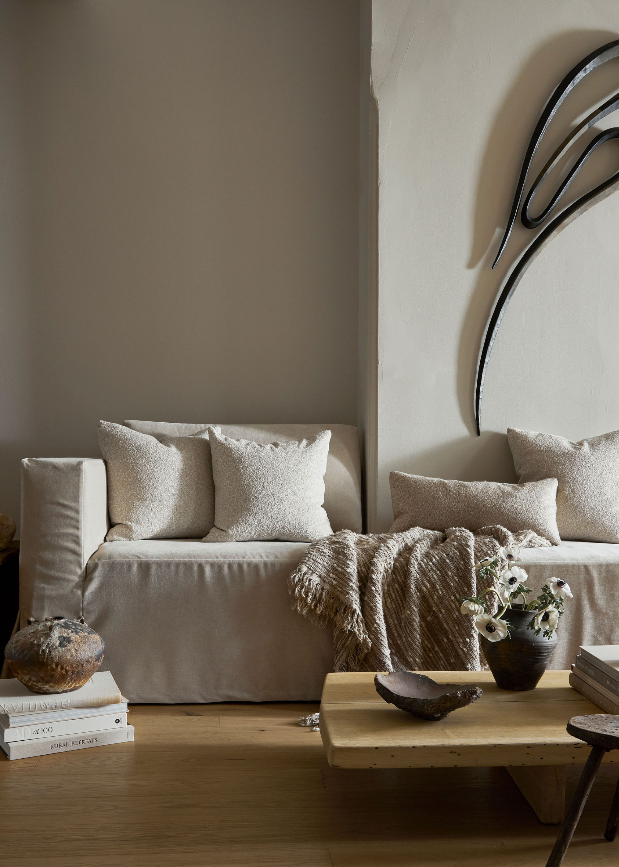 A living room with an off-white couch, a couple of pillows, a fringed throw, a vase, a stack of books, and a low wood coffee table with a vase of flowers