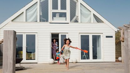 A little girl runs out of a beach house with a plastic shovel in her hand.