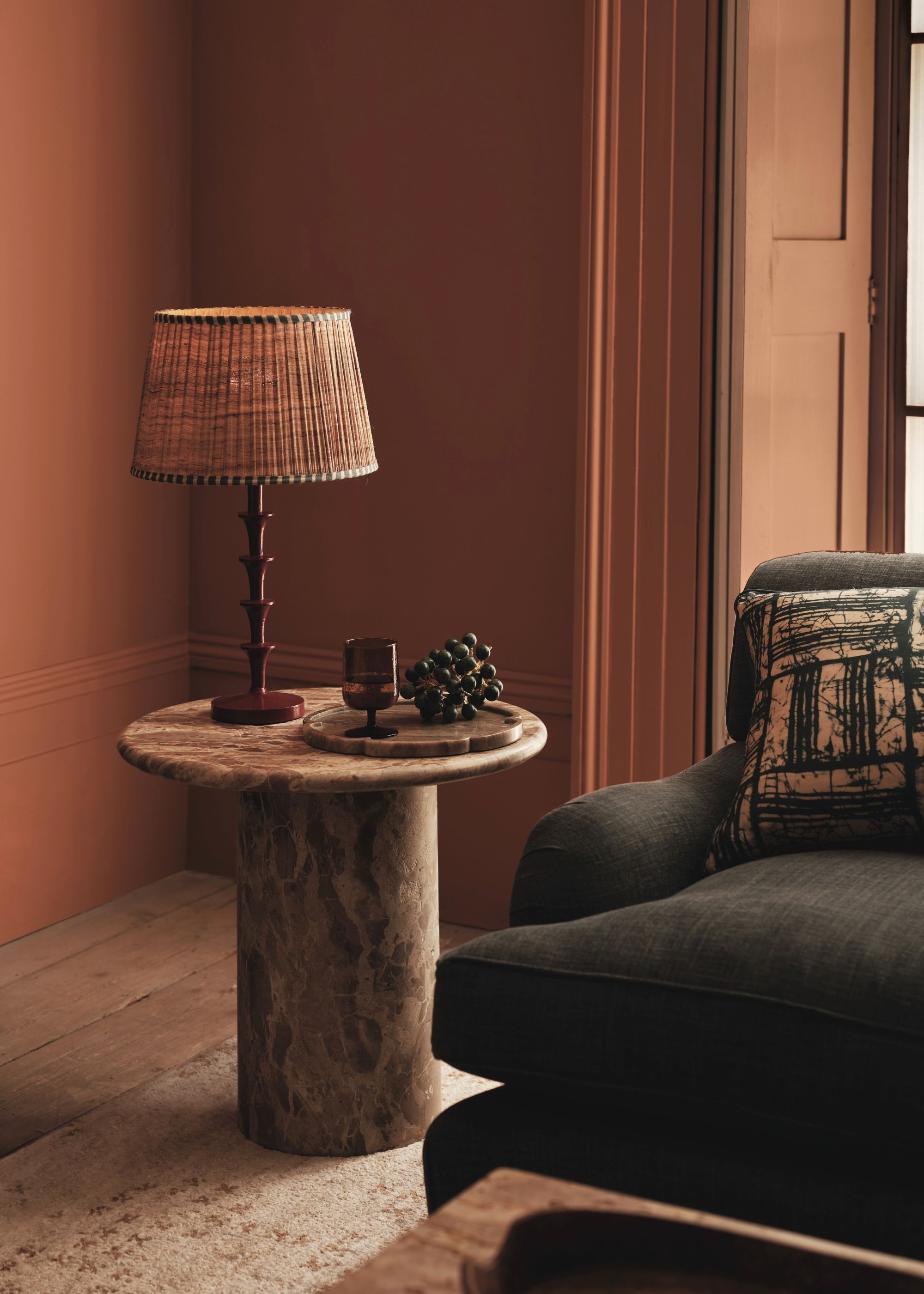 A stacked maroon colored table lamp with a lamp shade on a marble table next to a tray of grapes and a glass. There is also a view of the corner of a sofa with a cushion on top