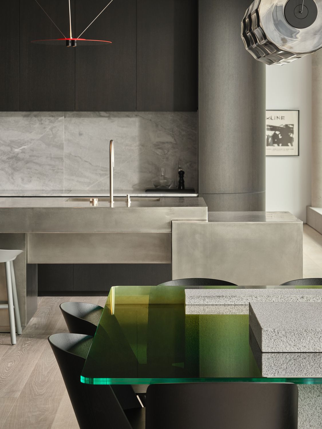 close up of dining table with green glass top with concrete pillars, stainless steel kitchen in background with dark cabinets and marble backsplash