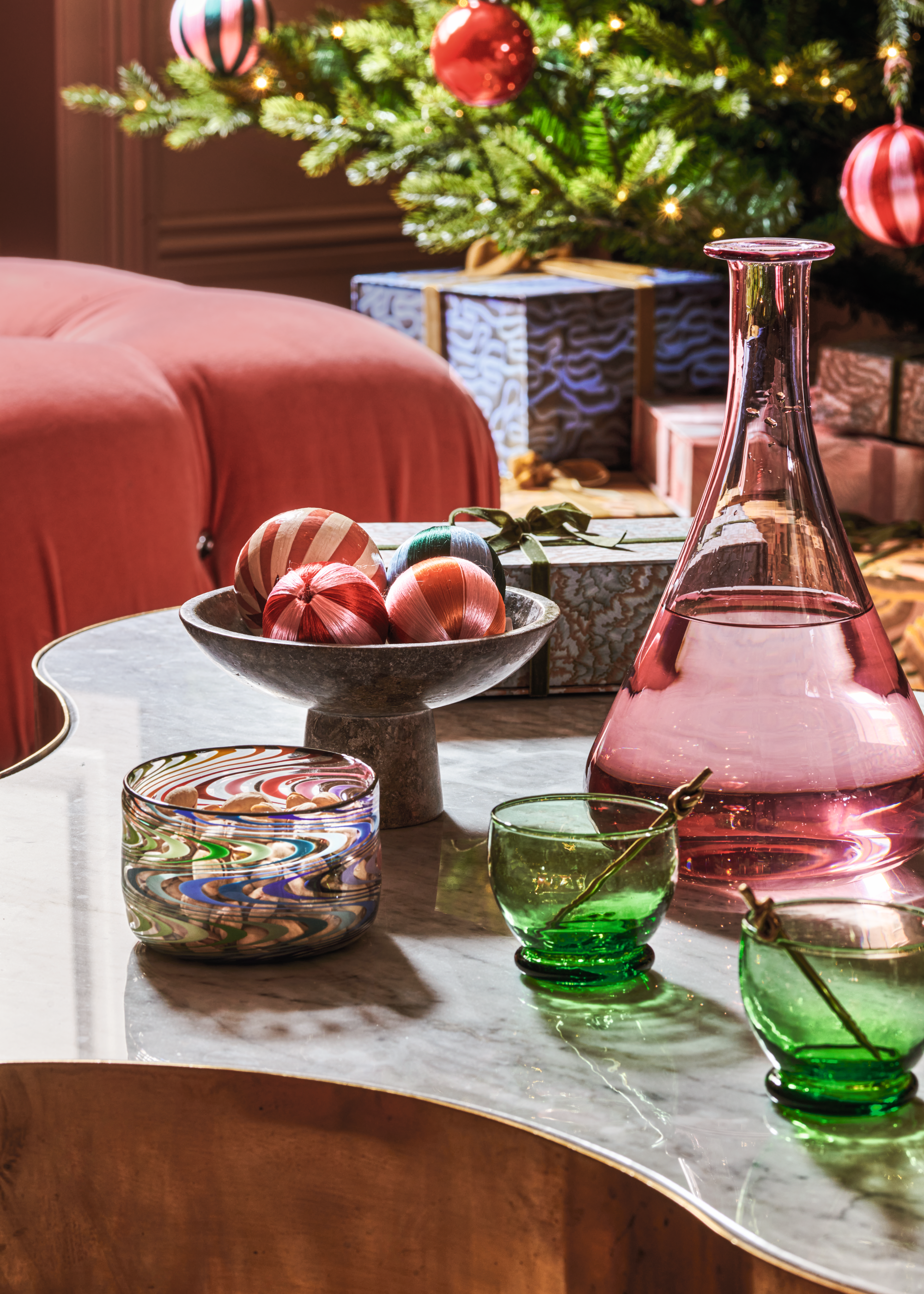 A coffee table with colorful glassware and a marble pedestal bowl with striped colorful baubles displayed inside and matching the decorations on the tree in the background