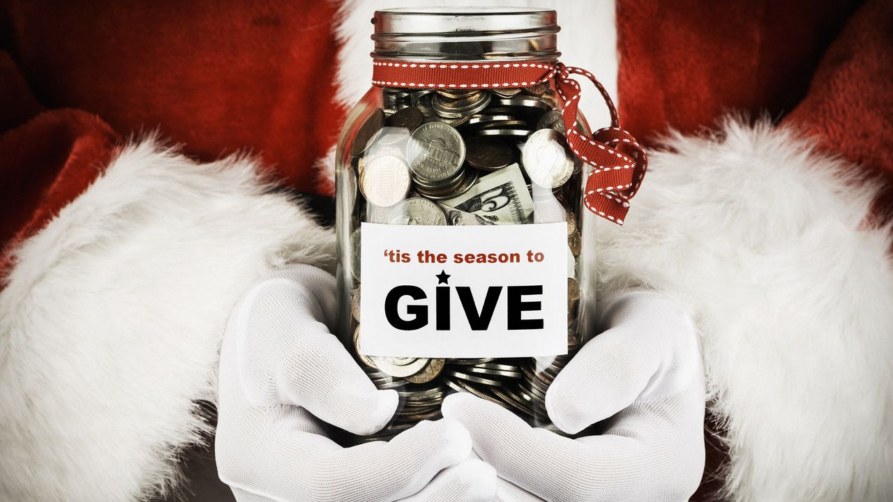 A person dressed in a Santa suit holds a jar filled with money and labeled &#039;Tis the Season to Give.