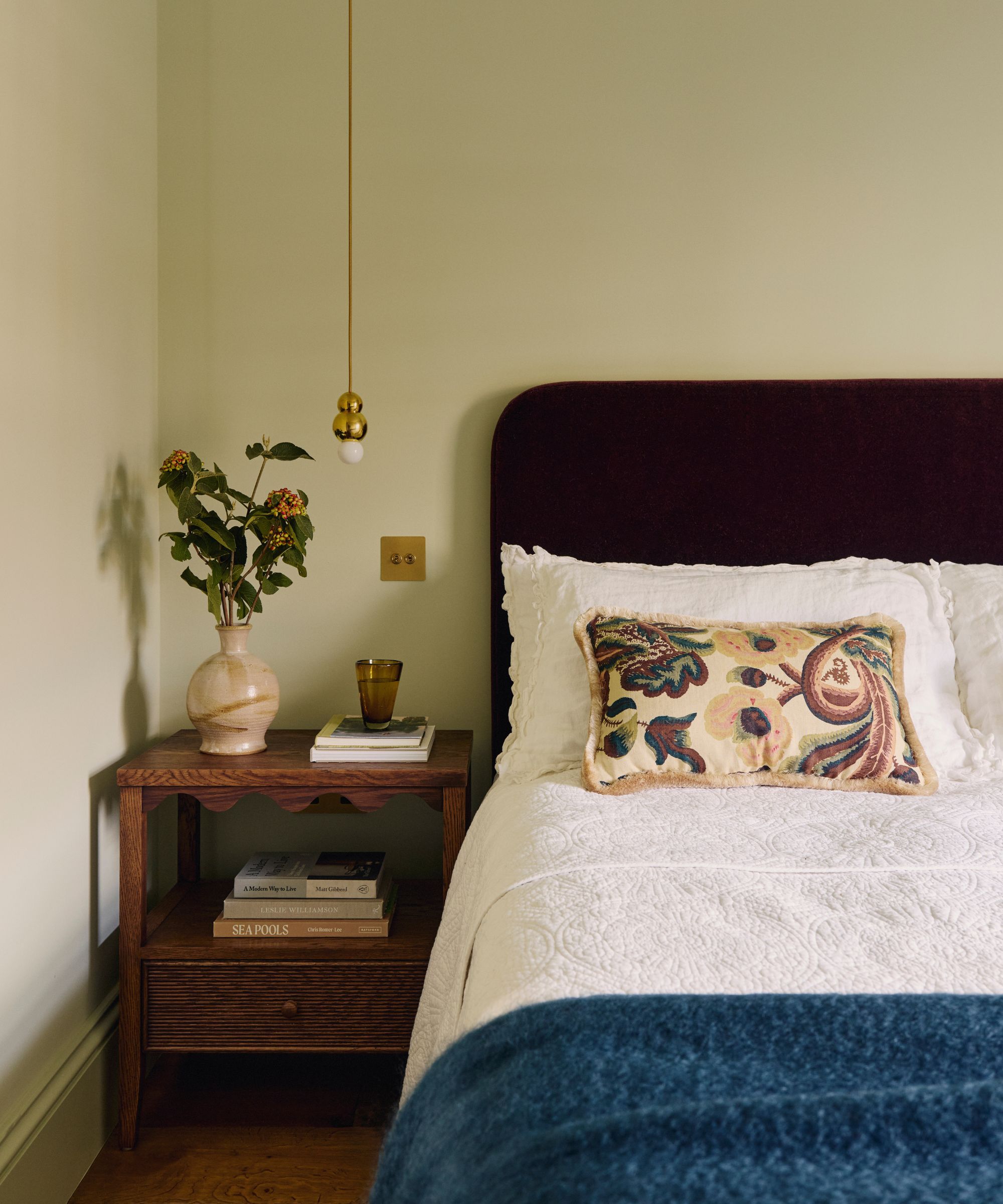 Bedroom with pale sage walls, a dark upholstered velvet bed, white bedding, a blue wool throw, and a tapestry cushion, with a wooden scalloped nightstand beside it decorated with books and a vase.