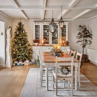 a dining room decorated for Christmas with sideboard and display case at one end of the table, a Christmas tree in the corner and white painted rustic dining furniture