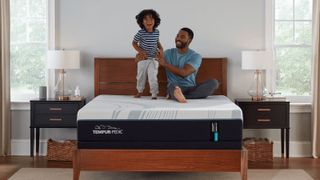 The Tempur-Pedic Tempur-Adapt mattress on a bed frame in a bedroom, a man sits on top of the mattress holding onto his young child, who is standing on the mattress