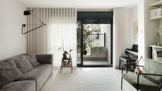 A living room with half open curtains and an open door leading to a balcony. In the living room is a large sofa and a chair opposite it. A large carpet sits in the middle of the room as well as a piano in the corner of the room