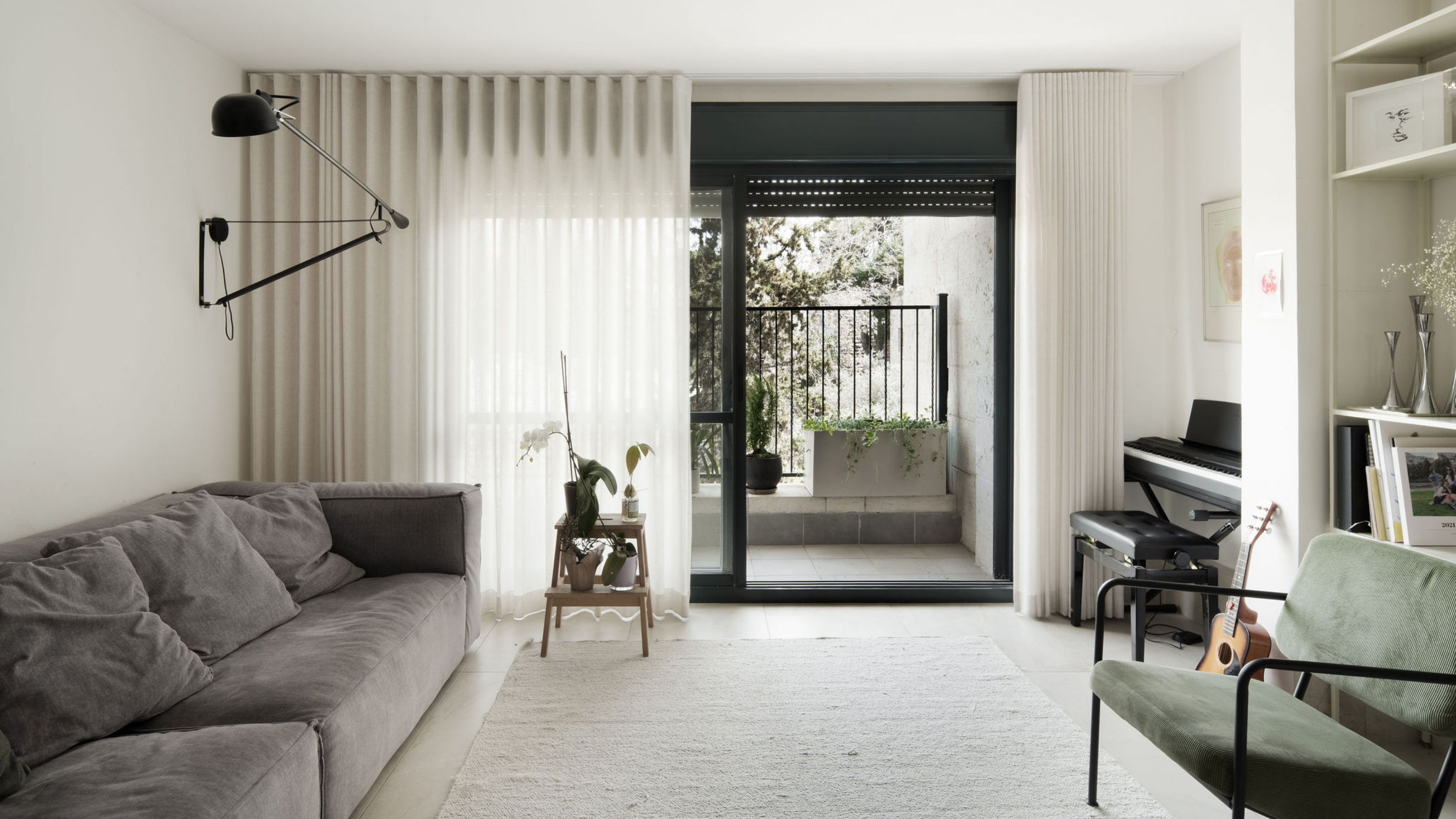 A living room with half open curtains and an open door leading to a balcony. In the living room is a large sofa and a chair opposite it. A large carpet sits in the middle of the room as well as a piano in the corner of the room 
