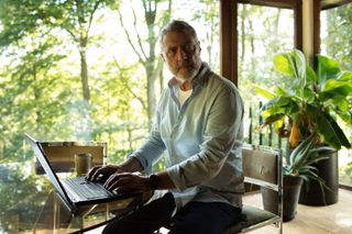 Billy (Vincent Regan) is sitting at a laptop in a room with floor-to-ceiling windows and woodland in view behind him